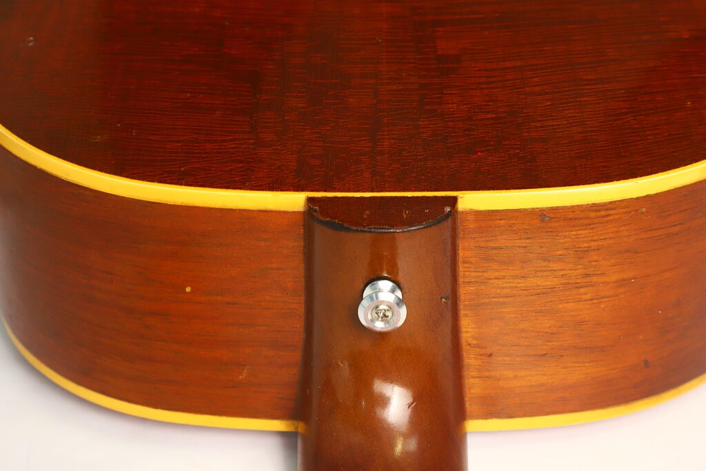 Nahaufnahme der Rückseite einer Gibson LG-3 Akustikgitarre aus dem Jahr 1954, mit Fokus auf den Halsansatz und einen glänzenden Gurtknopf, der dort angebracht ist, wo der Hals auf den Korpus trifft. Das Holz ist rotbraun lackiert und mit einer kontrastierenden gelben Einfassung versehen. Nahaufnahme der Rückseite einer Gibson LG-3 Akustikgitarre aus dem Jahr 1954, mit Fokus auf den Halsansatz und einen glänzenden Gurtknopf, der dort angebracht ist, wo der Hals auf den Korpus trifft. Das Holz ist rotbraun lackiert und mit einer kontrastierenden gelben Einfassung versehen.