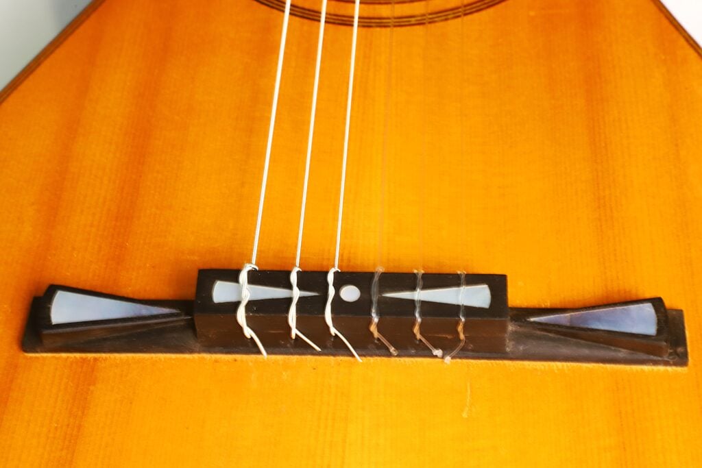 Nahaufnahme des Stegs und der Saiten einer Antonio Garcia de Lorca 1847 Replika Klassische Gitarre, die die feinen Details ihrer Bauweise und der Saitenbefestigung hervorhebt. Nahaufnahme des Stegs und der Saiten einer Antonio Garcia de Lorca 1847 Replika Klassische Gitarre, die die feinen Details ihrer Bauweise und der Saitenbefestigung hervorhebt.