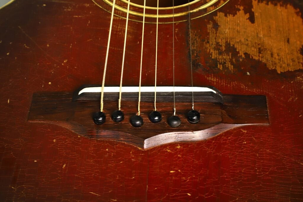 Nahaufnahme des Stegs und der Saiten einer Epiphone Recording B 1931 Akustikgitarre, auf der sichtbare Kratzer, Risse und eine verblasste Holzoberfläche zu erkennen sind. Nahaufnahme des Stegs und der Saiten einer Epiphone Recording B 1931 Akustikgitarre, auf der sichtbare Kratzer, Risse und eine verblasste Holzoberfläche zu erkennen sind.