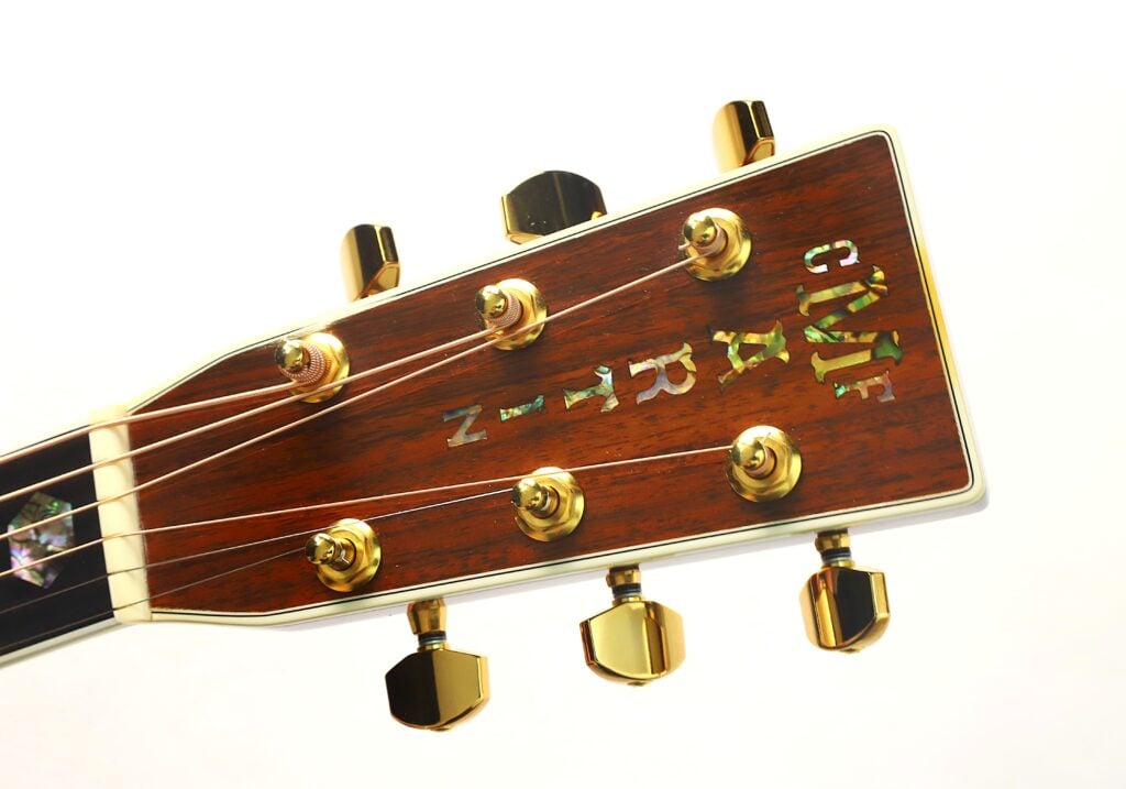Nahaufnahme des Kopfes einer Martin D-45 Akustikgitarre aus dem Jahr 2009 mit goldenen Stimmwirbeln und einer Inlay-Beschriftung aus Abalone auf einer Holzoberfläche, fotografiert vor weißem Hintergrund. Nahaufnahme des Kopfes einer Martin D-45 Akustikgitarre aus dem Jahr 2009 mit goldenen Stimmwirbeln und einer Inlay-Beschriftung aus Abalone auf einer Holzoberfläche, fotografiert vor weißem Hintergrund.