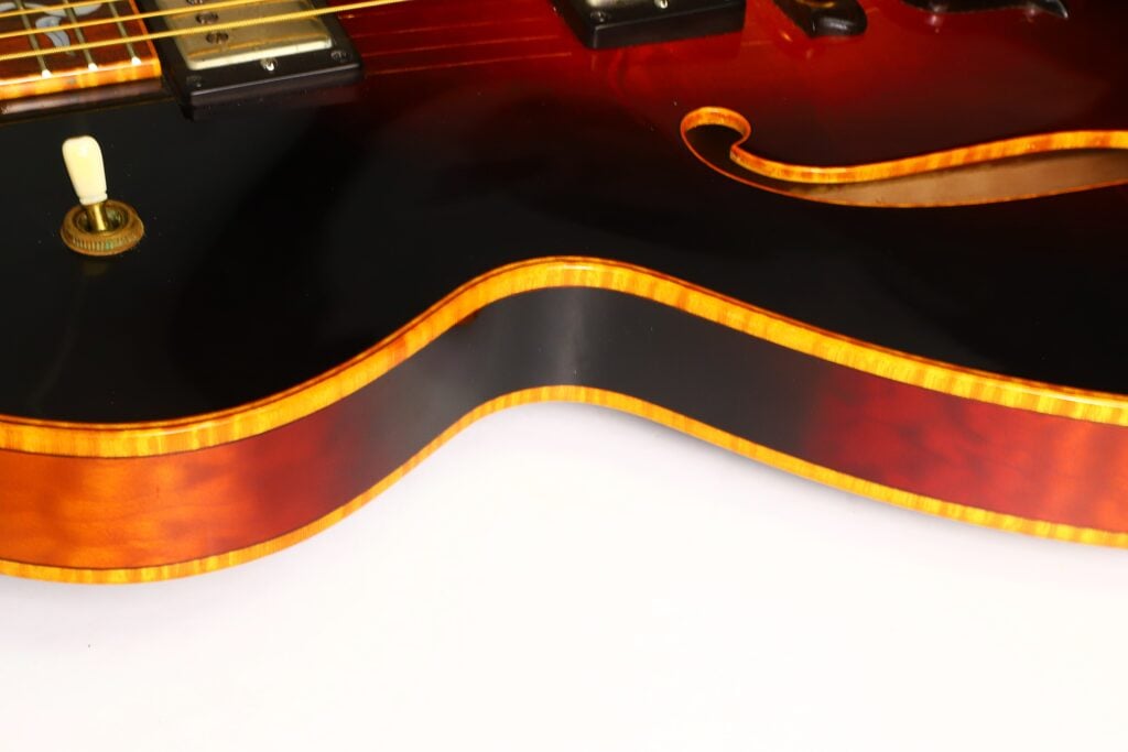 Nahaufnahme des Korpus der DeLaHaye 1995 Elektro-Akustik-Gitarre, die vor weißem Hintergrund ihre glänzende Sunburst-Lackierung, die orange-gelbe Einfassung, den Lautstärkeregler und die filigranen Details der F-Löcher zeigt. Nahaufnahme des Korpus der DeLaHaye 1995 Elektro-Akustik-Gitarre, die vor weißem Hintergrund ihre glänzende Sunburst-Lackierung, die orange-gelbe Einfassung, den Lautstärkeregler und die filigranen Details der F-Löcher zeigt.