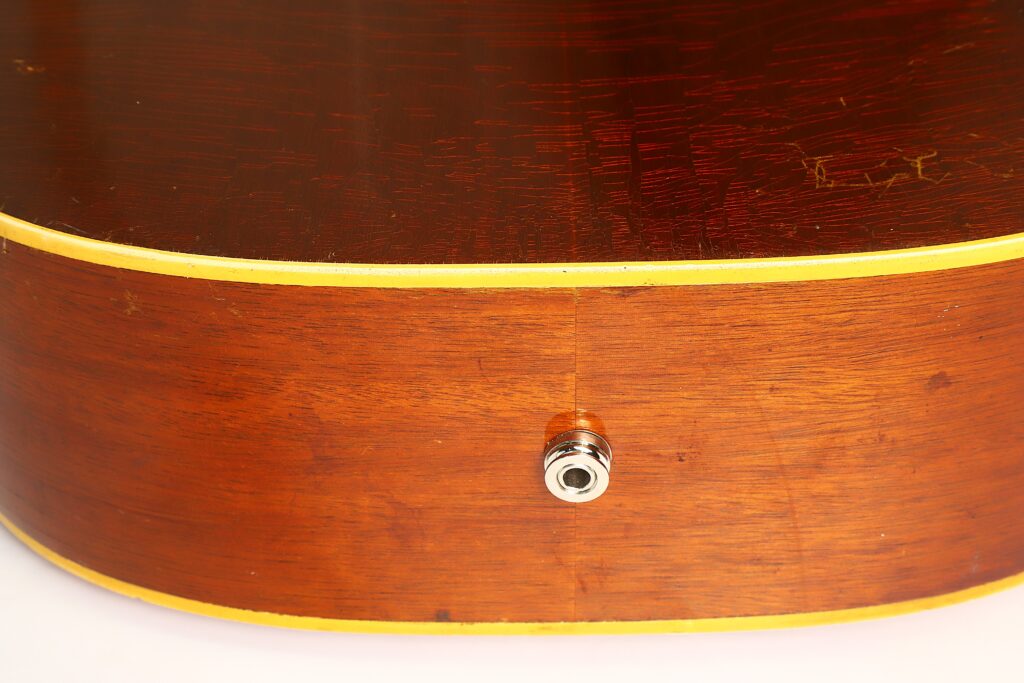 Nahaufnahme der Unterkante einer Gibson LG-3 Akustikgitarre aus dem Jahr 1954, die eine glänzende Metall-Eingangsbuchse zeigt, die in den Holzkorpus eingebaut ist, der entlang der Kante mit einer gelben Einfassung versehen ist. Nahaufnahme der Unterkante einer Gibson LG-3 Akustikgitarre aus dem Jahr 1954, die eine glänzende Metall-Eingangsbuchse zeigt, die in den Holzkorpus eingebaut ist, der entlang der Kante mit einer gelben Einfassung versehen ist.
