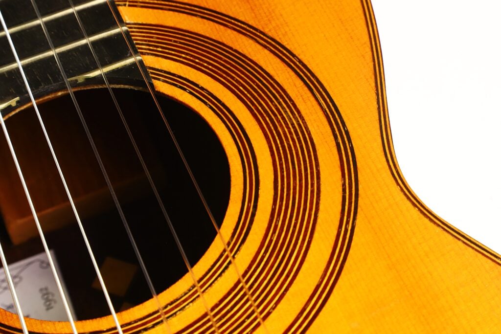 Nahaufnahme des Schalllochs und der Saiten der klassischen Gitarre „Antonio Garcia de Lorca 1847 Replika“, die die Holzmaserung, das kreisförmige Rosettenmuster und einen Teil des Instrumentenkörpers vor einem weißen Hintergrund hervorhebt. Nahaufnahme des Schalllochs und der Saiten der klassischen Gitarre „Antonio Garcia de Lorca 1847 Replika“, die die Holzmaserung, das kreisförmige Rosettenmuster und einen Teil des Instrumentenkörpers vor einem weißen Hintergrund hervorhebt.