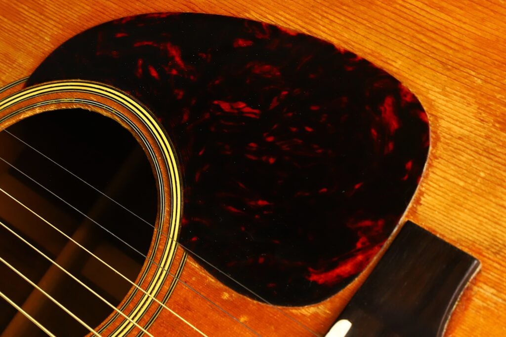 Nahaufnahme einer Martin D-28 Akustikgitarre aus dem Jahr 1965, auf der die Saiten, das Schallloch, das dunkle Schlagbrett mit Schildpatt-Muster und ein Teil des Holzkörpers zu sehen sind. Nahaufnahme einer Martin D-28 Akustikgitarre aus dem Jahr 1965, auf der die Saiten, das Schallloch, das dunkle Schlagbrett mit Schildpatt-Muster und ein Teil des Holzkörpers zu sehen sind.