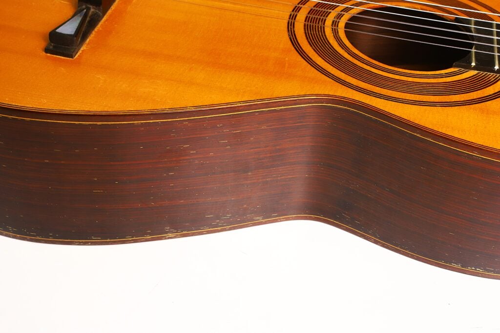 Nahaufnahme des Korpus der „Antonio Garcia de Lorca 1847 Replika Klassische Gitarre“, die die Holzmaserung hervorhebt; rechts sind vor weißem Hintergrund ein Teil der Saiten und das Schallloch zu sehen. Nahaufnahme des Korpus der „Antonio Garcia de Lorca 1847 Replika Klassische Gitarre“, die die Holzmaserung hervorhebt; rechts sind vor weißem Hintergrund ein Teil der Saiten und das Schallloch zu sehen.