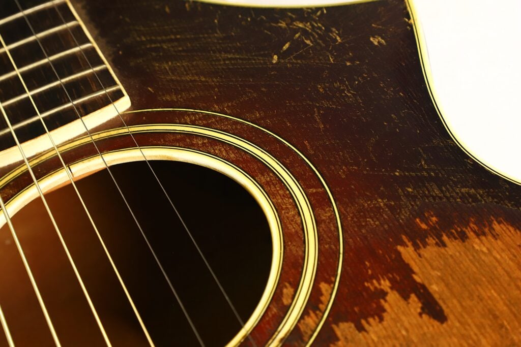 Nahaufnahme des Korpus und des Schalllochs einer Epiphone Recording B 1931 Akustikgitarre, auf der die Saiten sowie die abgenutzte, zerkratzte Holzoberfläche zu sehen sind. Nahaufnahme des Korpus und des Schalllochs einer Epiphone Recording B 1931 Akustikgitarre, auf der die Saiten sowie die abgenutzte, zerkratzte Holzoberfläche zu sehen sind.