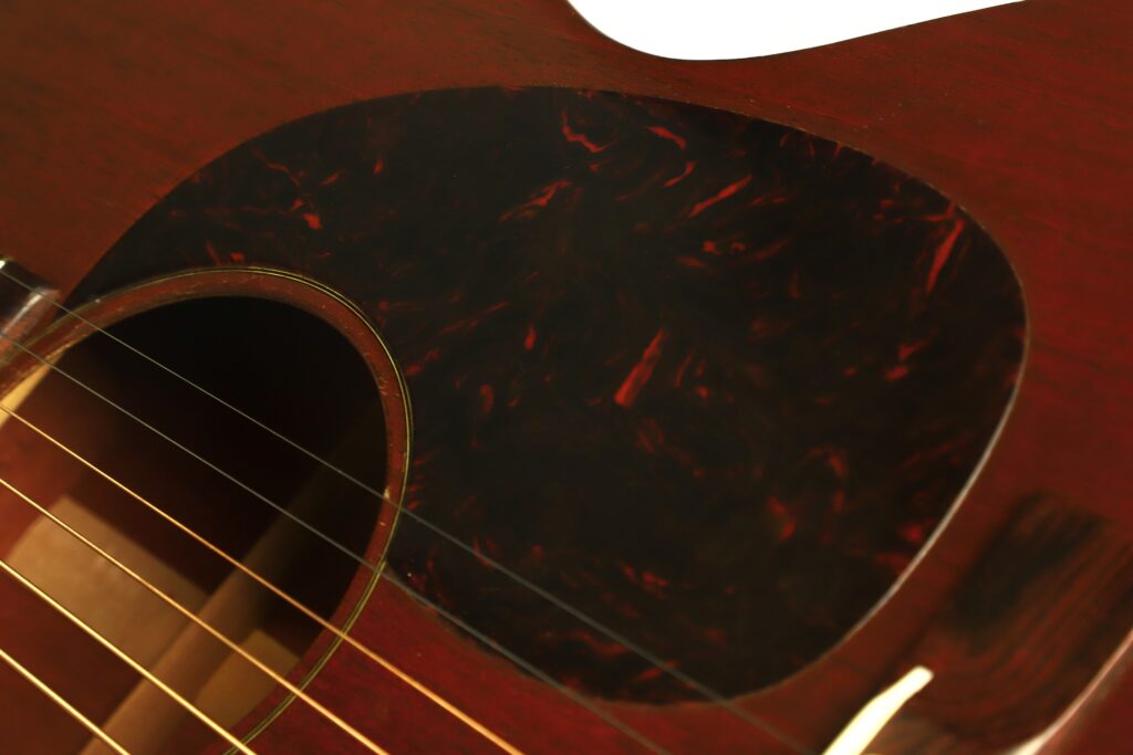 Nahaufnahme des Schalllochs, der Saiten und des Plektrumschutzes aus Schildpatt der Martin 00-17 Akustikgitarre von 1941, die die Holzmaserung und die feinen Details des Instruments hervorhebt. Nahaufnahme des Schalllochs, der Saiten und des Plektrumschutzes aus Schildpatt der Martin 00-17 Akustikgitarre von 1941, die die Holzmaserung und die feinen Details des Instruments hervorhebt.