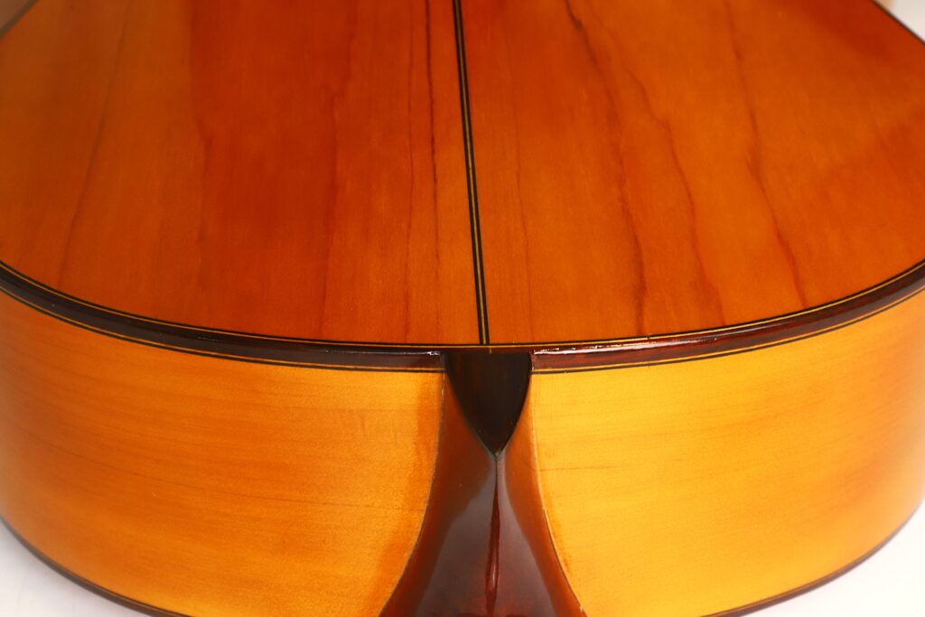 Nahaufnahme der Rückseite und der Zargen der Flamenco-Gitarre „Casa Arcangel Fernandez 1970“, die glänzendes, poliertes Holz mit sichtbarer Maserung und einer dunklen Mittelnaht zeigt. Nahaufnahme der Rückseite und der Zargen der Flamenco-Gitarre „Casa Arcangel Fernandez 1970“, die glänzendes, poliertes Holz mit sichtbarer Maserung und einer dunklen Mittelnaht zeigt.
