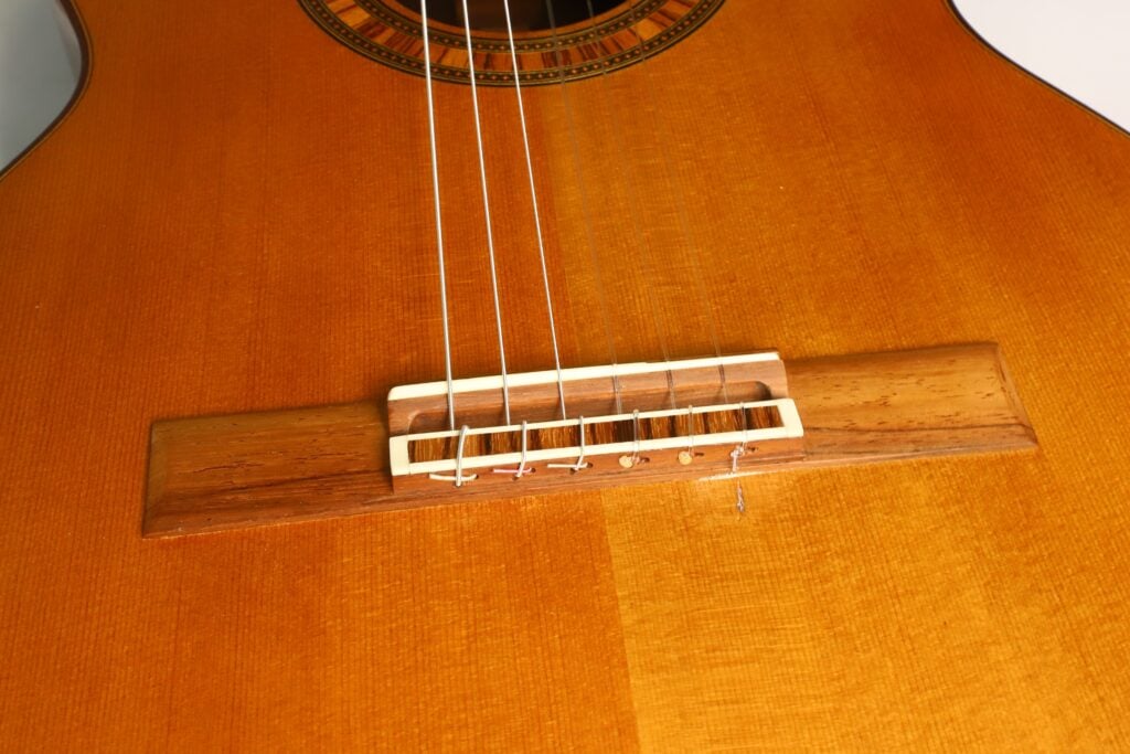 Nahaufnahme des Stegs und der Saiten der „Tosca Klassische Gitarre“ von Frank-Peter Dietrich aus dem Jahr 1998 auf ihrem Holzkorpus, wobei die detaillierte Holzmaserung, der Steg und der Sattel besonders hervorgehoben werden. Nahaufnahme des Stegs und der Saiten der „Tosca Klassische Gitarre“ von Frank-Peter Dietrich aus dem Jahr 1998 auf ihrem Holzkorpus, wobei die detaillierte Holzmaserung, der Steg und der Sattel besonders hervorgehoben werden.
