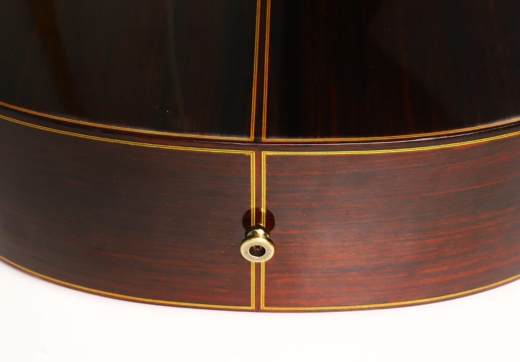 Nahaufnahme der Rückseite und der Seite einer Paulino Bernabe M-5 2008 Klassische Gitarre, die die glänzende, dunkle Holzmaserung, die gelben Einlegelinien und einen Metall-Gurtknopf unten in der Mitte hervorhebt. Nahaufnahme der Rückseite und der Seite einer Paulino Bernabe M-5 2008 Klassische Gitarre, die die glänzende, dunkle Holzmaserung, die gelben Einlegelinien und einen Metall-Gurtknopf unten in der Mitte hervorhebt.