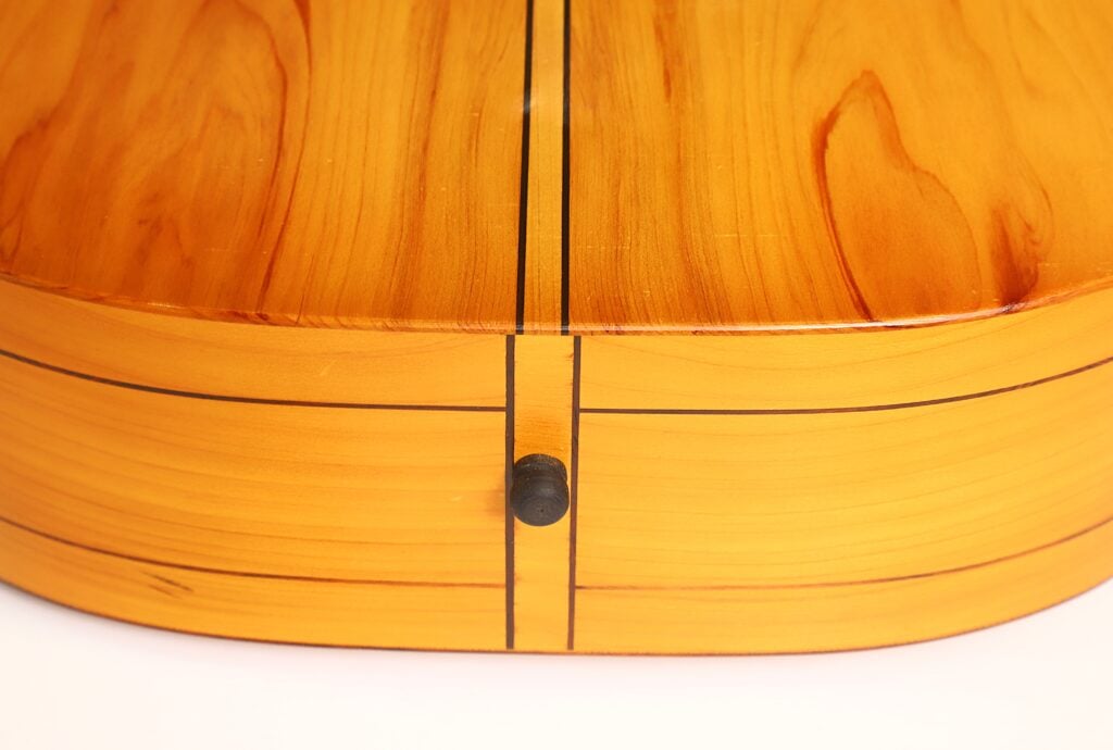 Nahaufnahme der Rückseite einer Vihuela von Hans Herb aus dem Jahr 1989, die poliertes Holz, schwarze Linien, die die Form umreißen, und einen Riemenknopf unten in der Mitte zeigt. Nahaufnahme der Rückseite einer Vihuela von Hans Herb aus dem Jahr 1989, die poliertes Holz, schwarze Linien, die die Form umreißen, und einen Riemenknopf unten in der Mitte zeigt.