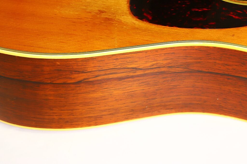 Nahaufnahme der Seite einer Martin D-28 Akustikgitarre aus dem Jahr 1965, die die Holzmaserung und die glänzende Lackierung zeigt; dabei sind ein Teil der Decke und das Schlagbrett der Gitarre zu sehen. Nahaufnahme der Seite einer Martin D-28 Akustikgitarre aus dem Jahr 1965, die die Holzmaserung und die glänzende Lackierung zeigt; dabei sind ein Teil der Decke und das Schlagbrett der Gitarre zu sehen.
