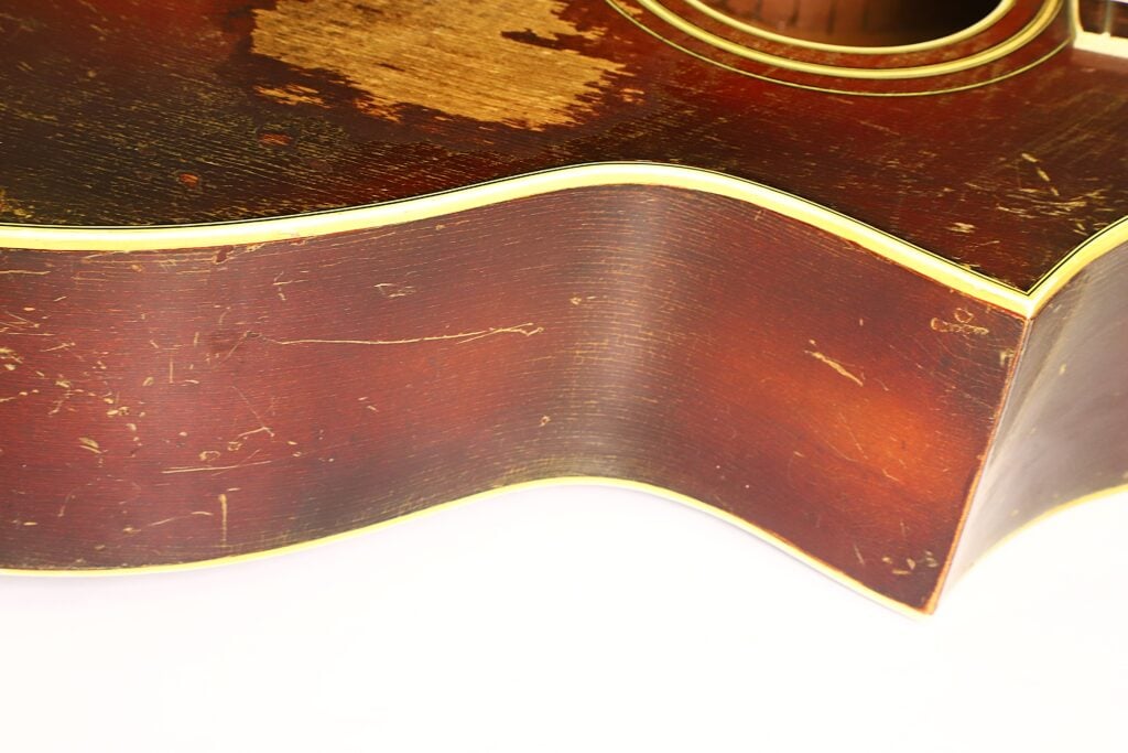 Nahaufnahme der Seite einer abgenutzten Epiphone Recording B 1931 Vintage-Akustikgitarre mit sichtbaren Kratzern, abgeplatztem Holz und gelber Einfassung vor weißem Hintergrund. Nahaufnahme der Seite einer abgenutzten Epiphone Recording B 1931 Vintage-Akustikgitarre mit sichtbaren Kratzern, abgeplatztem Holz und gelber Einfassung vor weißem Hintergrund.