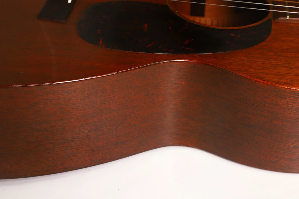 Nahaufnahme der Seite und des unteren Teils einer Martin 00-17 Akustikgitarre aus dem Jahr 1941, die die Maserung des Holzes und die sanften Rundungen hervorhebt. Die Gitarre hat eine glänzende braune Lackierung und liegt auf einer weißen Unterlage. Nahaufnahme der Seite und des unteren Teils einer Martin 00-17 Akustikgitarre aus dem Jahr 1941, die die Maserung des Holzes und die sanften Rundungen hervorhebt. Die Gitarre hat eine glänzende braune Lackierung und liegt auf einer weißen Unterlage.
