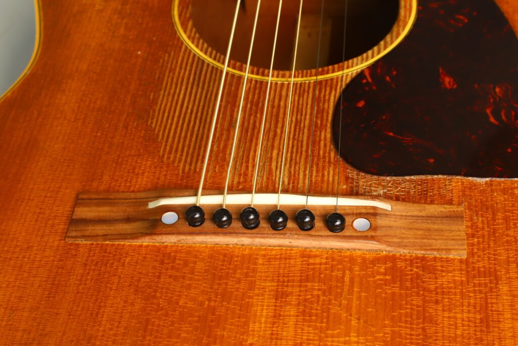 Nahaufnahme des Holzstegs der Gibson LG-3 Akustikgitarre von 1954 mit sechs Saiten und schwarzen Stiften, wobei die Maserung und Struktur des Holzes sowie ein Teil des dunklen Schlagbretts hervorgehoben werden. Nahaufnahme des Holzstegs der Gibson LG-3 Akustikgitarre von 1954 mit sechs Saiten und schwarzen Stiften, wobei die Maserung und Struktur des Holzes sowie ein Teil des dunklen Schlagbretts hervorgehoben werden.