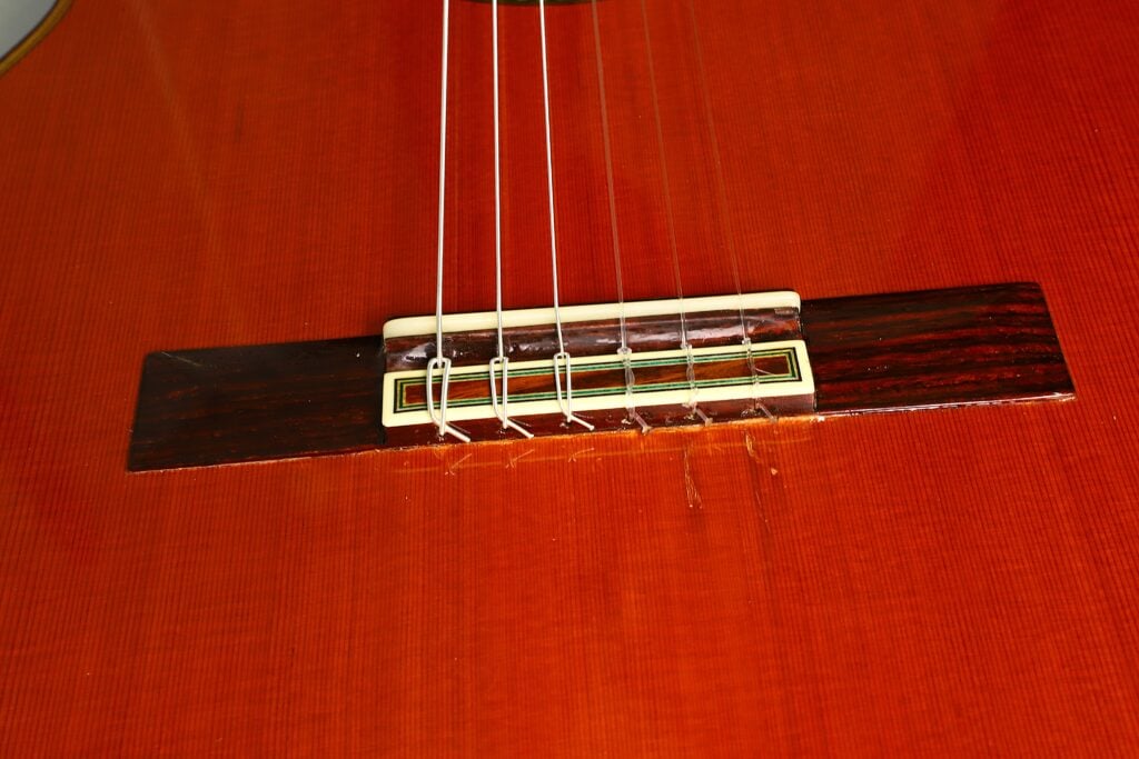 Nahaufnahme des Stegs und der Saiten einer Paulino Bernabe M-5 2008 Klassische Gitarre, die den polierten Holzkorpus, die feine Maserung und das aufwendige Design im Bereich des Stegs hervorhebt. Nahaufnahme des Stegs und der Saiten einer Paulino Bernabe M-5 2008 Klassische Gitarre, die den polierten Holzkorpus, die feine Maserung und das aufwendige Design im Bereich des Stegs hervorhebt.
