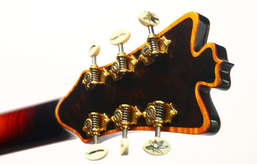 Nahaufnahme der Rückseite des Kopfes einer DeLaHaye Elektro-Akustik-Gitarre aus dem Jahr 1995, die sechs Stimmwirbel mit goldfarbenen Beschlägen und ein markantes, kantiges Holzdesign mit glänzender Oberfläche zeigt, ergänzt durch weiße Stimmknöpfe mit dekorativen Einlagen. Nahaufnahme der Rückseite des Kopfes einer DeLaHaye Elektro-Akustik-Gitarre aus dem Jahr 1995, die sechs Stimmwirbel mit goldfarbenen Beschlägen und ein markantes, kantiges Holzdesign mit glänzender Oberfläche zeigt, ergänzt durch weiße Stimmknöpfe mit dekorativen Einlagen.