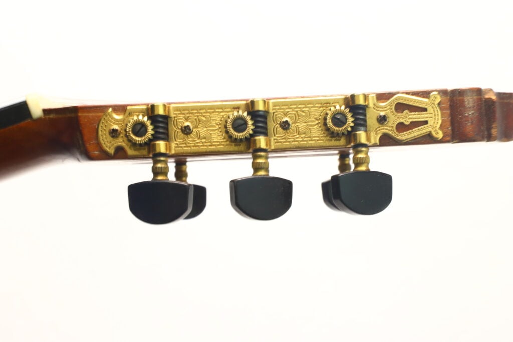 Seitenansicht des Kopfes der „Andres Martin 1966 Klassische Gitarre“ mit drei schwarzen Stimmwirbeln und kunstvoll verzierten goldenen Mechaniken vor weißem Hintergrund. Seitenansicht des Kopfes der „Andres Martin 1966 Klassische Gitarre“ mit drei schwarzen Stimmwirbeln und kunstvoll verzierten goldenen Mechaniken vor weißem Hintergrund.