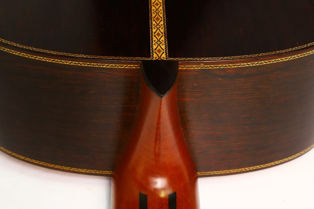 Nahaufnahme der Rückseite der „Masaru Kohno Modell 50 1979 Klassische Gitarre“, an der der Hals auf den elegant verzierten Korpus aus dunklem Holz trifft, der an den Kanten mit aufwendigen Einlegearbeiten verziert ist. Nahaufnahme der Rückseite der „Masaru Kohno Modell 50 1979 Klassische Gitarre“, an der der Hals auf den elegant verzierten Korpus aus dunklem Holz trifft, der an den Kanten mit aufwendigen Einlegearbeiten verziert ist.