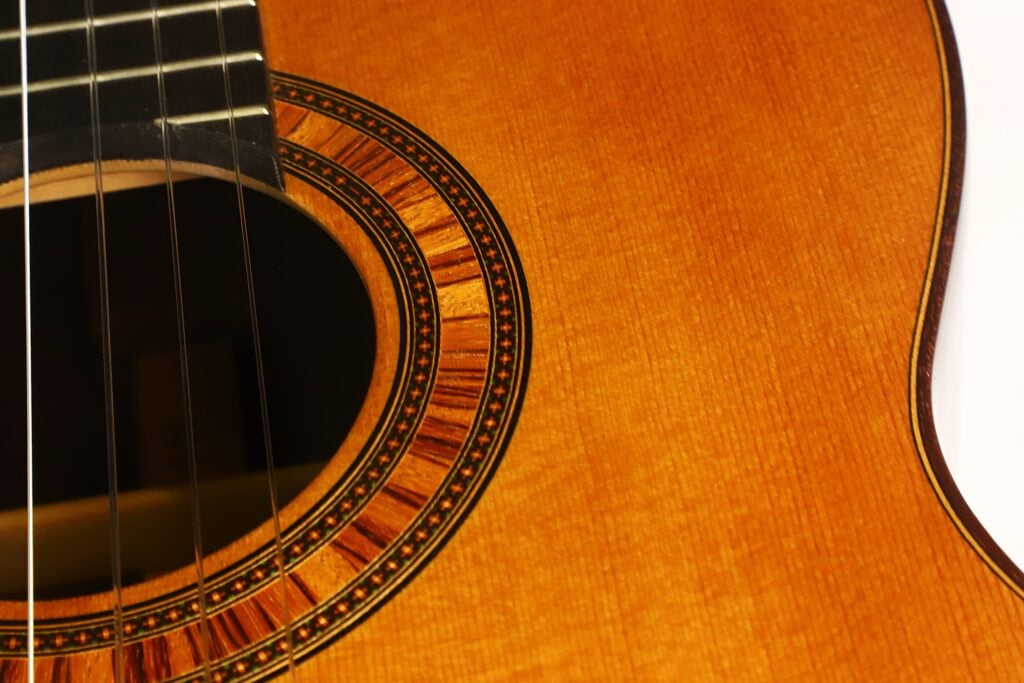 Eine Nahaufnahme der „Tosca Klassische Gitarre“ von Frank-Peter Dietrich aus dem Jahr 1998 zeigt das Schallloch und die Saiten mit ihrer filigranen Holzmaserung sowie eine elegante Zierrosette, die die Öffnung umgibt. Eine Nahaufnahme der „Tosca Klassische Gitarre“ von Frank-Peter Dietrich aus dem Jahr 1998 zeigt das Schallloch und die Saiten mit ihrer filigranen Holzmaserung sowie eine elegante Zierrosette, die die Öffnung umgibt.