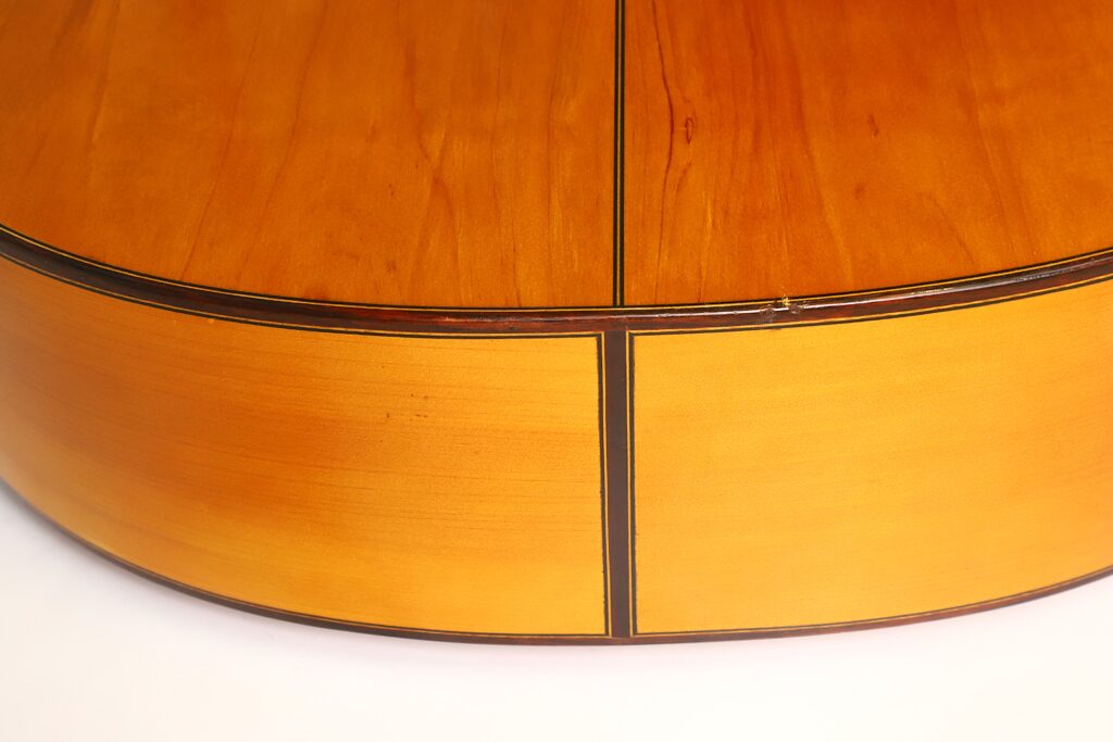 Nahaufnahme der Rückseite und der Seite der Flamenco-Gitarre „Casa Arcangel Fernandez 1970“, die die feine Holzmaserung, die glänzende Lackierung und die dekorativen dunklen Zierlinien an den Fugen der Platten hervorhebt. Nahaufnahme der Rückseite und der Seite der Flamenco-Gitarre „Casa Arcangel Fernandez 1970“, die die feine Holzmaserung, die glänzende Lackierung und die dekorativen dunklen Zierlinien an den Fugen der Platten hervorhebt.