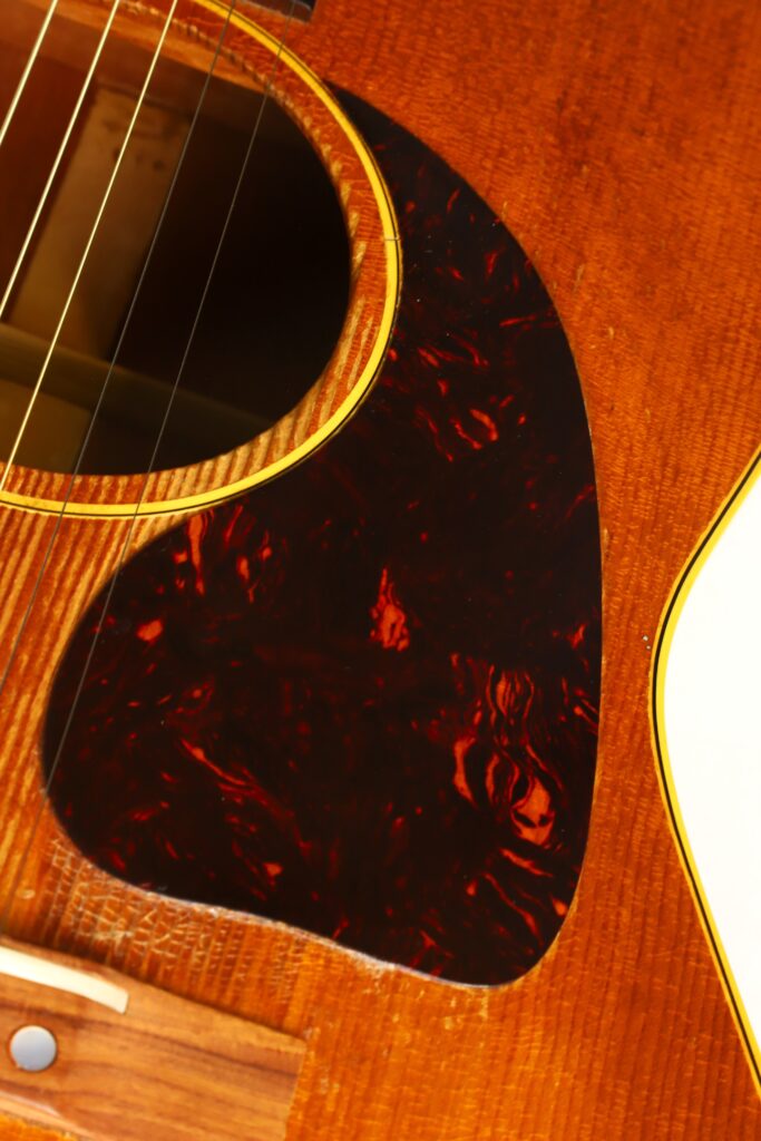 Nahaufnahme einer Gibson LG-3 Akustikgitarre aus dem Jahr 1954, die das Schallloch, das rot marmorierte Schlagbrett sowie einen Teil des Holzkörpers und des Stegs zeigt. Nahaufnahme einer Gibson LG-3 Akustikgitarre aus dem Jahr 1954, die das Schallloch, das rot marmorierte Schlagbrett sowie einen Teil des Holzkörpers und des Stegs zeigt.