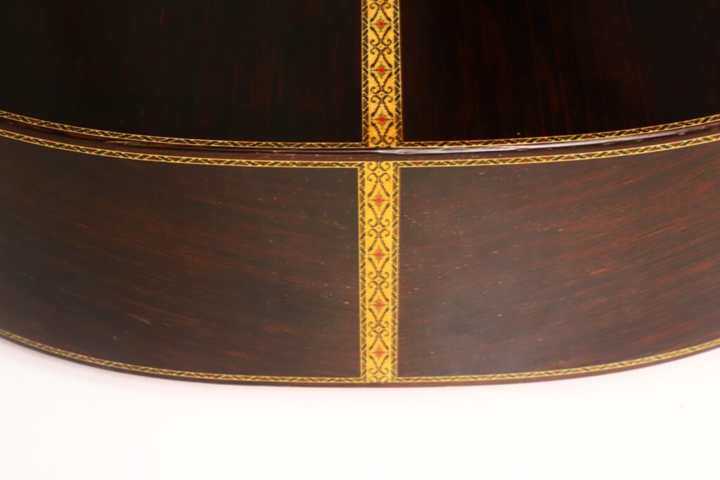 Nahaufnahme der klassischen Gitarre „Masaru Kohno Modell 50, 1979“ mit einem dunklen Holzkorpus, der mit dekorativen gelben und roten Einlagen verziert ist, die vertikal und horizontal verlaufen und auf der glatten, geschwungenen Oberfläche ein Kreuzmuster bilden. Nahaufnahme der klassischen Gitarre „Masaru Kohno Modell 50, 1979“ mit einem dunklen Holzkorpus, der mit dekorativen gelben und roten Einlagen verziert ist, die vertikal und horizontal verlaufen und auf der glatten, geschwungenen Oberfläche ein Kreuzmuster bilden.