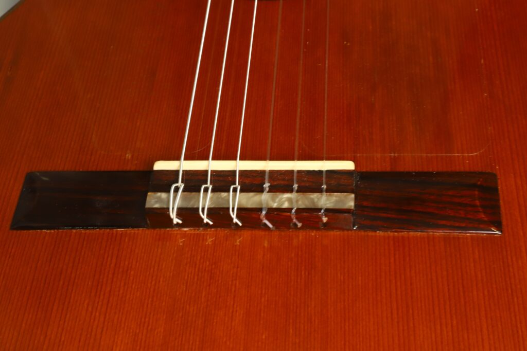 Nahaufnahme des Stegs einer Flamenco-Gitarre von Casa Arcangel Fernandez aus dem Jahr 1970, deren Nylonsaiten am rotbraunen Holzkorpus festgebunden sind. Das Bild hebt die feinen Maserungsdetails des Holzes hervor. Nahaufnahme des Stegs einer Flamenco-Gitarre von Casa Arcangel Fernandez aus dem Jahr 1970, deren Nylonsaiten am rotbraunen Holzkorpus festgebunden sind. Das Bild hebt die feinen Maserungsdetails des Holzes hervor.