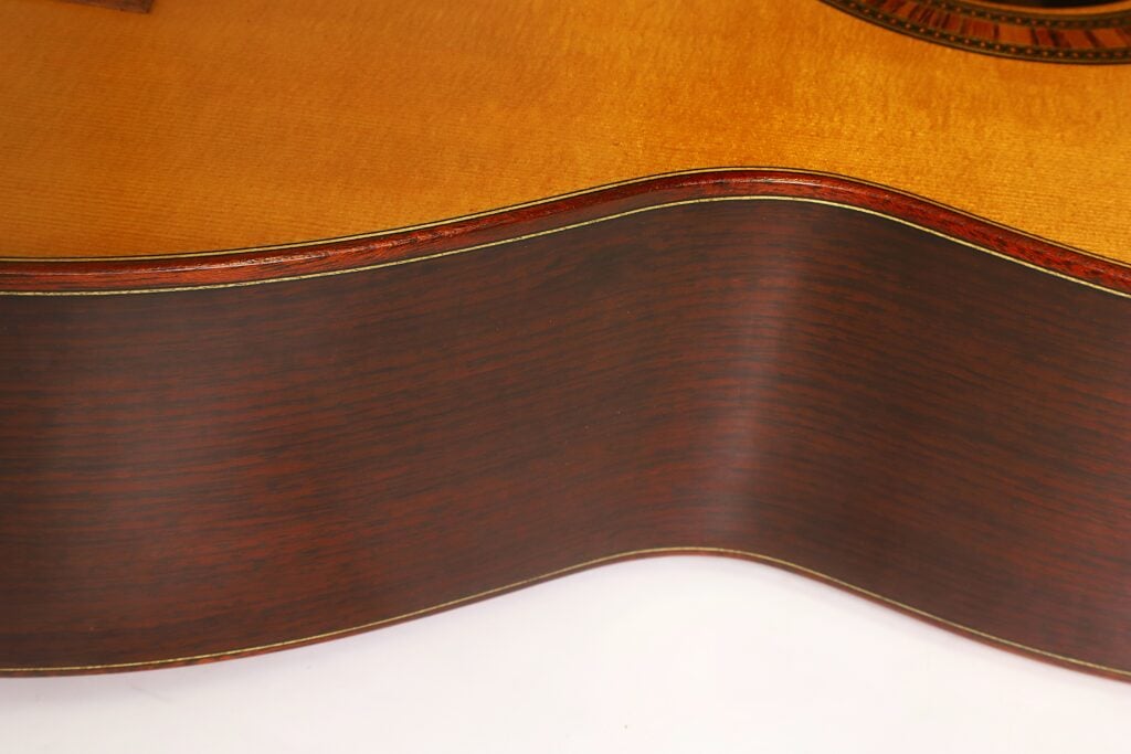 Nahaufnahme der Seite der „Tosca Klassische Gitarre“ von Frank-Peter Dietrich aus dem Jahr 1998, die vor weißem Hintergrund die dunkle Holzmaserung und die geschwungene Silhouette hervorhebt. Nahaufnahme der Seite der „Tosca Klassische Gitarre“ von Frank-Peter Dietrich aus dem Jahr 1998, die vor weißem Hintergrund die dunkle Holzmaserung und die geschwungene Silhouette hervorhebt.