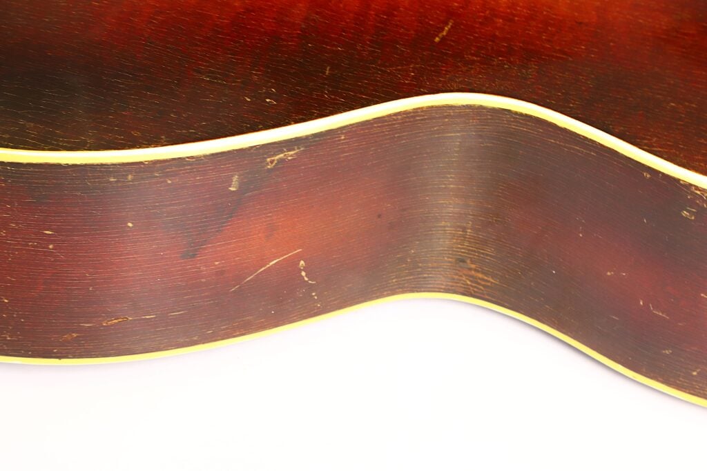 Nahaufnahme der Seite einer Epiphone Recording B 1931 Akustikgitarre mit rotbrauner Lackierung, sichtbarer Holzmaserung, leichten Kratzern und cremefarbener Einfassung vor weißem Hintergrund. Nahaufnahme der Seite einer Epiphone Recording B 1931 Akustikgitarre mit rotbrauner Lackierung, sichtbarer Holzmaserung, leichten Kratzern und cremefarbener Einfassung vor weißem Hintergrund.