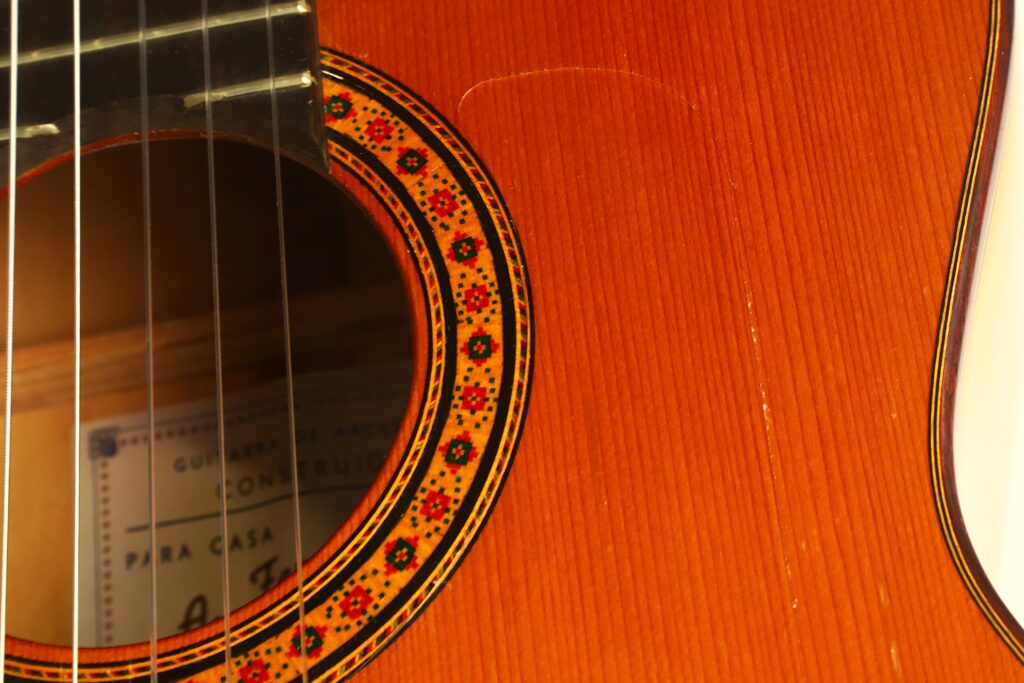 Nahaufnahme des Korpus einer Flamenco-Gitarre der Casa Arcangel Fernandez aus dem Jahr 1970, bei der das Schallloch, das aufwendige Rosettenmuster und die markante Holzmaserung besonders hervorgehoben sind. Durch das Schallloch ist zudem ein Teil des Innenetiketts zu sehen. Nahaufnahme des Korpus einer Flamenco-Gitarre der Casa Arcangel Fernandez aus dem Jahr 1970, bei der das Schallloch, das aufwendige Rosettenmuster und die markante Holzmaserung besonders hervorgehoben sind. Durch das Schallloch ist zudem ein Teil des Innenetiketts zu sehen.