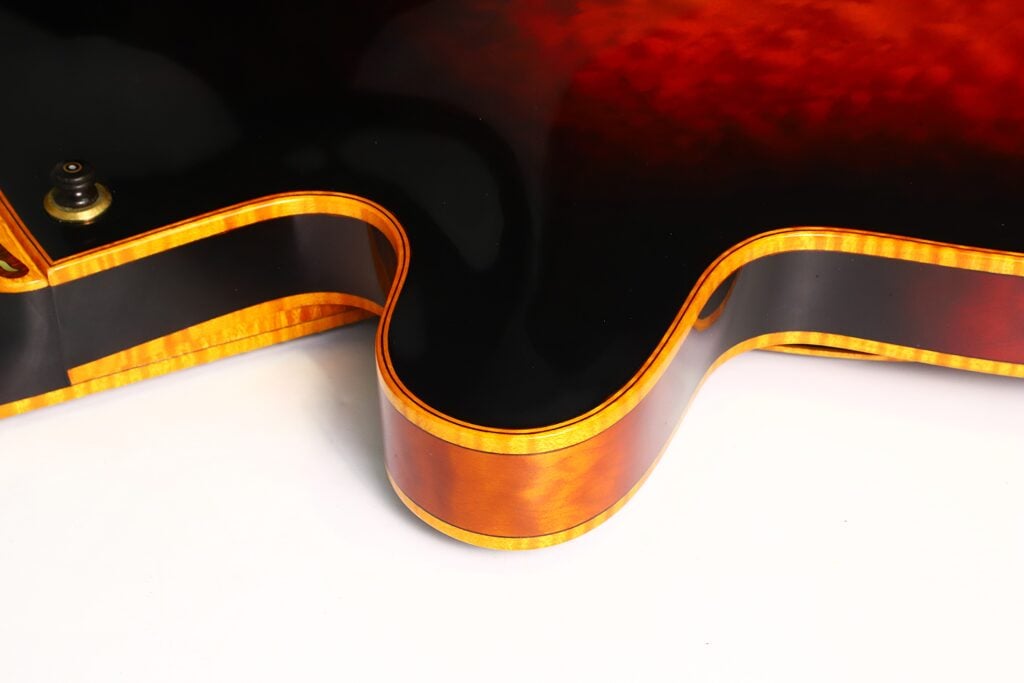 Nahaufnahme des Korpus der DeLaHaye 1995 Elektro-Akustik-Gitarre, die ihre Sunburst-Lackierung mit einer glänzend schwarzen Decke und leuchtend orangefarbenen Zargen hervorhebt. Das Bild konzentriert sich auf die geschwungene Kante und die detailreiche Einfassung der Gitarre vor einem weißen Hintergrund. Nahaufnahme des Korpus der DeLaHaye 1995 Elektro-Akustik-Gitarre, die ihre Sunburst-Lackierung mit einer glänzend schwarzen Decke und leuchtend orangefarbenen Zargen hervorhebt. Das Bild konzentriert sich auf die geschwungene Kante und die detailreiche Einfassung der Gitarre vor einem weißen Hintergrund.