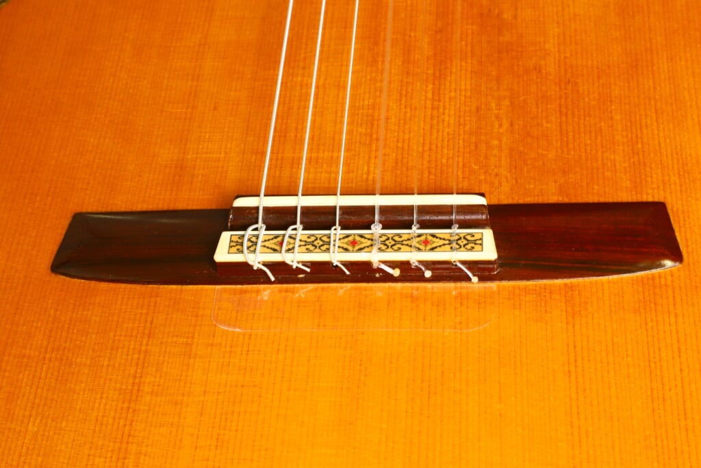 Nahaufnahme des Stegs einer klassischen Gitarre vom Typ „Masaru Kohno Modell 50“ aus dem Jahr 1979 mit dekorativen Einlagen und Nylonsaiten, die am Holzkorpus befestigt sind. Die Decke besticht durch eine warme, orange-braune Lackierung. Nahaufnahme des Stegs einer klassischen Gitarre vom Typ „Masaru Kohno Modell 50“ aus dem Jahr 1979 mit dekorativen Einlagen und Nylonsaiten, die am Holzkorpus befestigt sind. Die Decke besticht durch eine warme, orange-braune Lackierung.