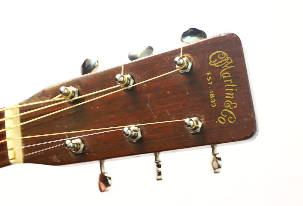 Nahaufnahme des Kopfes einer Martin 5-18 Akustikgitarre aus dem Jahr 1958, auf der sechs Stimmwirbel und das Martin & Co.-Logo mit der Aufschrift „Est. 1833“ auf der Holzoberfläche zu sehen sind, vor weißem Hintergrund. Nahaufnahme des Kopfes einer Martin 5-18 Akustikgitarre aus dem Jahr 1958, auf der sechs Stimmwirbel und das Martin & Co.-Logo mit der Aufschrift „Est. 1833“ auf der Holzoberfläche zu sehen sind, vor weißem Hintergrund.