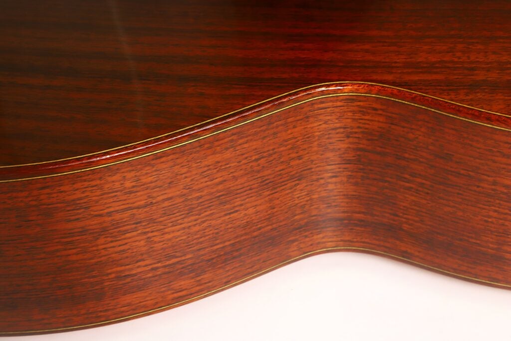 Detaillierte Nahaufnahme des Korpus der „Tosca Klassische Gitarre“ von Frank-Peter Dietrich aus dem Jahr 1998, die die geschwungenen Konturen, die feine Holzmaserung, die glatte, reflektierende Lackierung und die aufwendig gestalteten, mehrschichtigen Kantenverzierungen hervorhebt. Detaillierte Nahaufnahme des Korpus der „Tosca Klassische Gitarre“ von Frank-Peter Dietrich aus dem Jahr 1998, die die geschwungenen Konturen, die feine Holzmaserung, die glatte, reflektierende Lackierung und die aufwendig gestalteten, mehrschichtigen Kantenverzierungen hervorhebt.