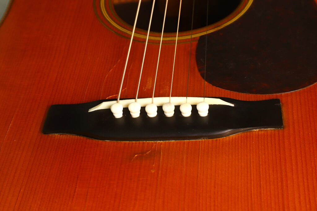 Nahaufnahme des Stegs und der Saiten einer Martin D-28, einer Akustikgitarre aus dem Jahr 1941 (Vor dem Krieg), auf der sechs Saiten zu sehen sind, die an weißen Stegstiften am Holzkorpus befestigt sind. Nahaufnahme des Stegs und der Saiten einer Martin D-28, einer Akustikgitarre aus dem Jahr 1941 (Vor dem Krieg), auf der sechs Saiten zu sehen sind, die an weißen Stegstiften am Holzkorpus befestigt sind.