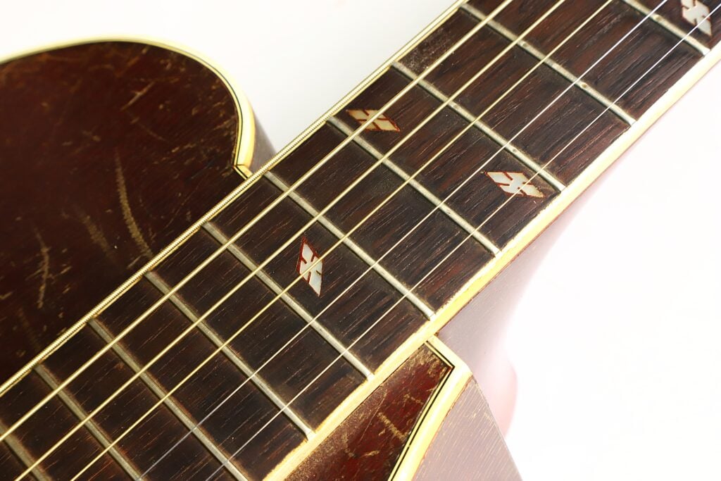 Nahaufnahme des braunen Halses und Korpus einer Epiphone Recording B 1931 Akustikgitarre mit vier metallischen Einlagen auf dem Griffbrett und einer gelben Zierleiste, die das Holz umrahmt. Die Oberfläche der Gitarre weist leichte Gebrauchsspuren auf. Nahaufnahme des braunen Halses und Korpus einer Epiphone Recording B 1931 Akustikgitarre mit vier metallischen Einlagen auf dem Griffbrett und einer gelben Zierleiste, die das Holz umrahmt. Die Oberfläche der Gitarre weist leichte Gebrauchsspuren auf.