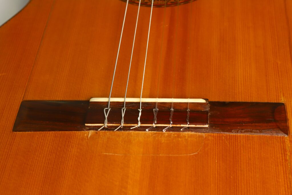 Nahaufnahme der Saiten und des Stegs einer Francisco-Barba-Flamenco-Gitarre aus dem Jahr 1966, die die Maserung des Holzes sowie die Details des Stegs und der Saitenbefestigungspunkte hervorhebt. Nahaufnahme der Saiten und des Stegs einer Francisco-Barba-Flamenco-Gitarre aus dem Jahr 1966, die die Maserung des Holzes sowie die Details des Stegs und der Saitenbefestigungspunkte hervorhebt.