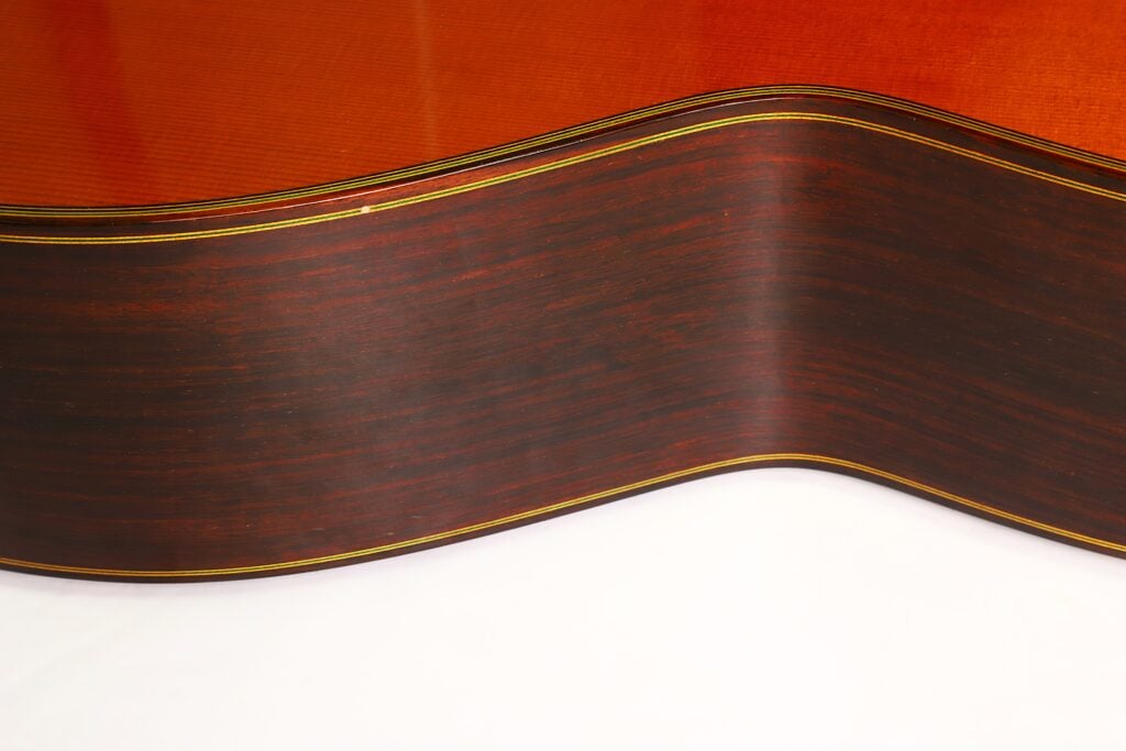 Nahaufnahme der dunklen Holzseite einer Paulino Bernabe M-5 2008 Klassische Gitarre, die ihre sanfte, wellenförmige Krümmung zeigt, während sie auf einer weißen Fläche ruht. Nahaufnahme der dunklen Holzseite einer Paulino Bernabe M-5 2008 Klassische Gitarre, die ihre sanfte, wellenförmige Krümmung zeigt, während sie auf einer weißen Fläche ruht.