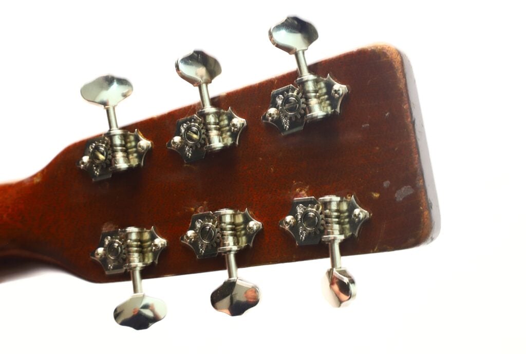 Nahaufnahme der Rückseite des Kopfes einer Martin 5-18 Akustikgitarre aus dem Jahr 1958, auf der sechs Metallstimmwirbel zu sehen sind, die auf einer braunen Holzfläche vor weißem Hintergrund angebracht sind. Nahaufnahme der Rückseite des Kopfes einer Martin 5-18 Akustikgitarre aus dem Jahr 1958, auf der sechs Metallstimmwirbel zu sehen sind, die auf einer braunen Holzfläche vor weißem Hintergrund angebracht sind.