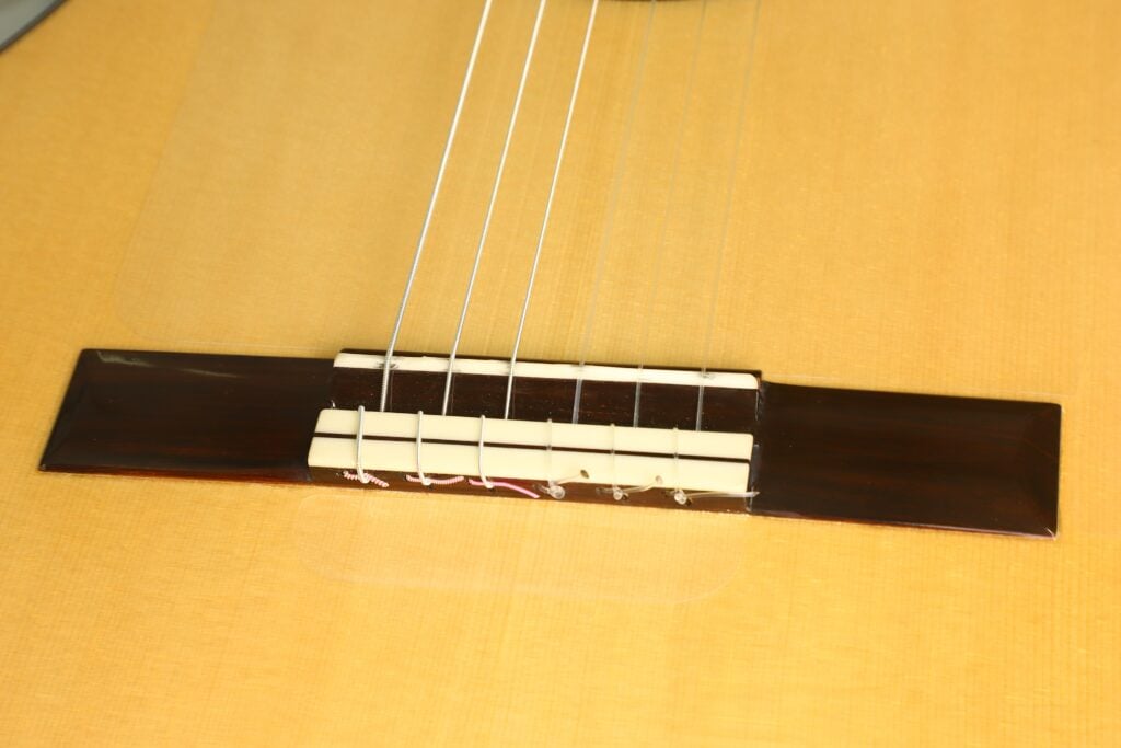 Nahaufnahme des Stegs einer Lester DeVoe 2020 Negra Flamenco-Gitarre, auf der die Saiten am Sattel eines Korpus aus hellem Holz befestigt sind. Nahaufnahme des Stegs einer Lester DeVoe 2020 Negra Flamenco-Gitarre, auf der die Saiten am Sattel eines Korpus aus hellem Holz befestigt sind.