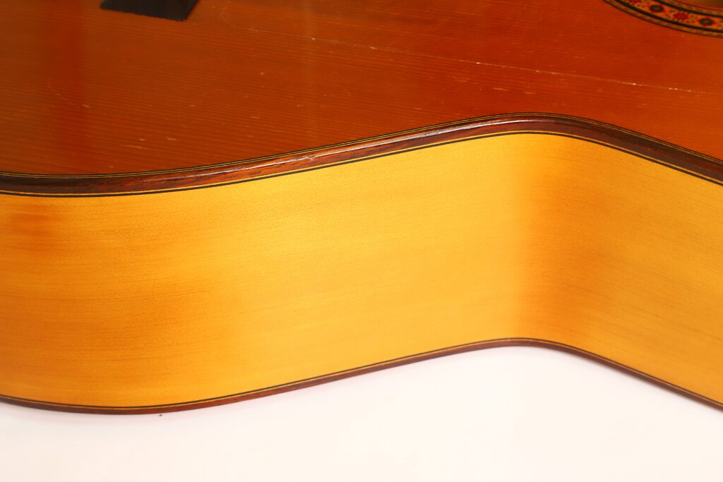 Nahaufnahme der Seite und des Korpus einer Flamenco-Gitarre von Casa Arcangel Fernandez aus dem Jahr 1970, die die Maserung des Holzes und die sanften Rundungen vor weißem Hintergrund hervorhebt. Nahaufnahme der Seite und des Korpus einer Flamenco-Gitarre von Casa Arcangel Fernandez aus dem Jahr 1970, die die Maserung des Holzes und die sanften Rundungen vor weißem Hintergrund hervorhebt.