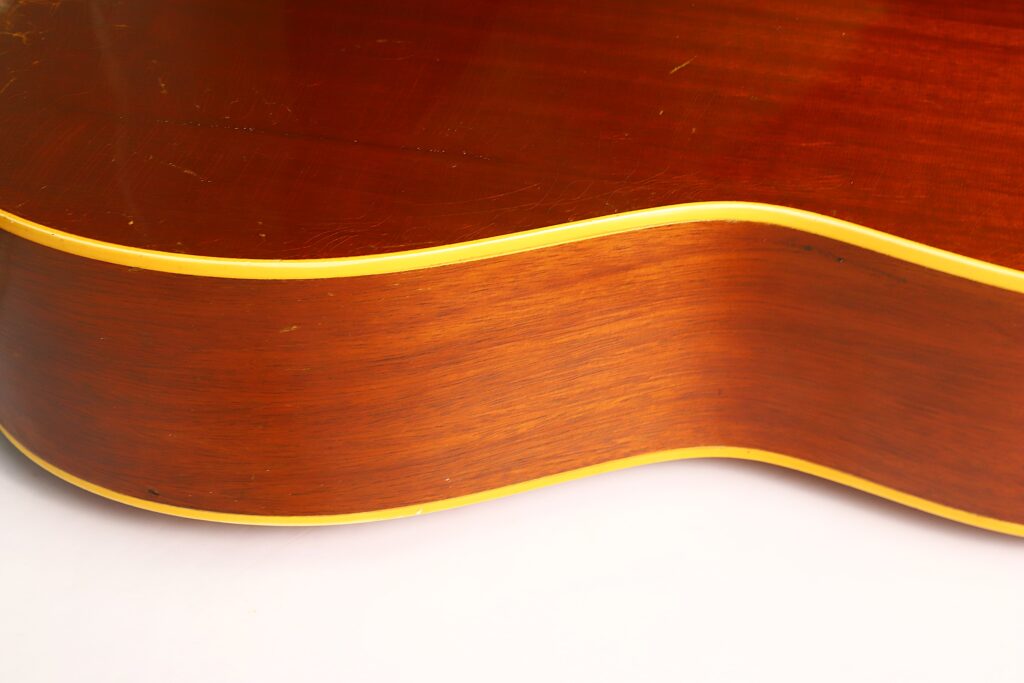 Nahaufnahme der Seite einer Gibson LG-3 Akustikgitarre aus dem Jahr 1954, die die Holzmaserung und die gelben Zierleisten vor weißem Hintergrund hervorhebt. Nahaufnahme der Seite einer Gibson LG-3 Akustikgitarre aus dem Jahr 1954, die die Holzmaserung und die gelben Zierleisten vor weißem Hintergrund hervorhebt.