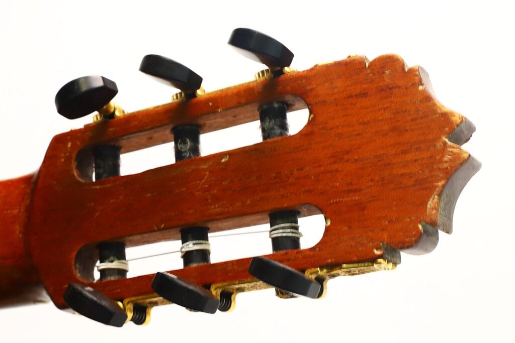 Eine detaillierte Nahaufnahme des hölzernen Kopfstücks der „Andres Martin 1966 Klassische Gitarre“ mit schwarzen Stimmwirbeln und Metallsaiten vor weißem Hintergrund. Eine detaillierte Nahaufnahme des hölzernen Kopfstücks der „Andres Martin 1966 Klassische Gitarre“ mit schwarzen Stimmwirbeln und Metallsaiten vor weißem Hintergrund.
