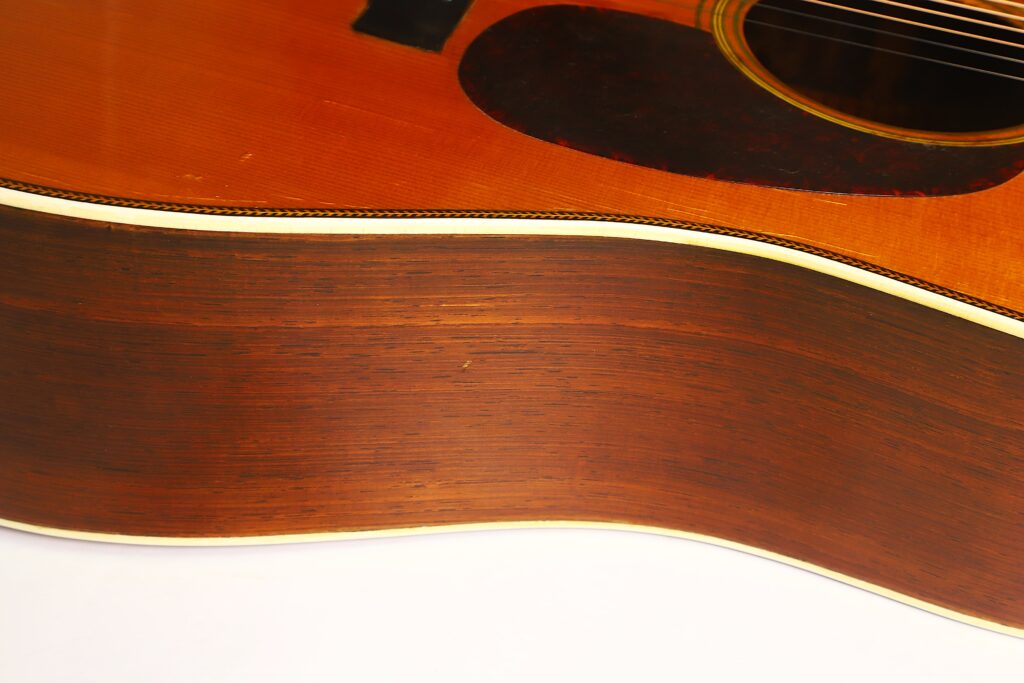 Nahaufnahme der Seite und des Korpus der Martin D-28 1941 Pre-War Akustikgitarre, die die Holzmaserung und die weiße Einfassung hervorhebt; oben rechts sind ein Teil des Schalllochs und das schwarze Schlagbrett zu sehen. Nahaufnahme der Seite und des Korpus der Martin D-28 1941 Pre-War Akustikgitarre, die die Holzmaserung und die weiße Einfassung hervorhebt; oben rechts sind ein Teil des Schalllochs und das schwarze Schlagbrett zu sehen.