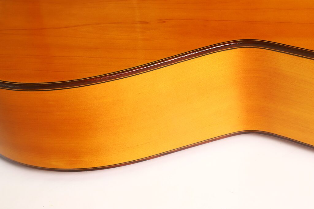 Nahaufnahme der Seite und Rückseite einer Flamenco-Gitarre von Casa Arcangel Fernandez aus dem Jahr 1970, die die glatte, helle Holzoberfläche und die dunkle Kantenverzierung vor einem schlichten weißen Hintergrund hervorhebt. Nahaufnahme der Seite und Rückseite einer Flamenco-Gitarre von Casa Arcangel Fernandez aus dem Jahr 1970, die die glatte, helle Holzoberfläche und die dunkle Kantenverzierung vor einem schlichten weißen Hintergrund hervorhebt.