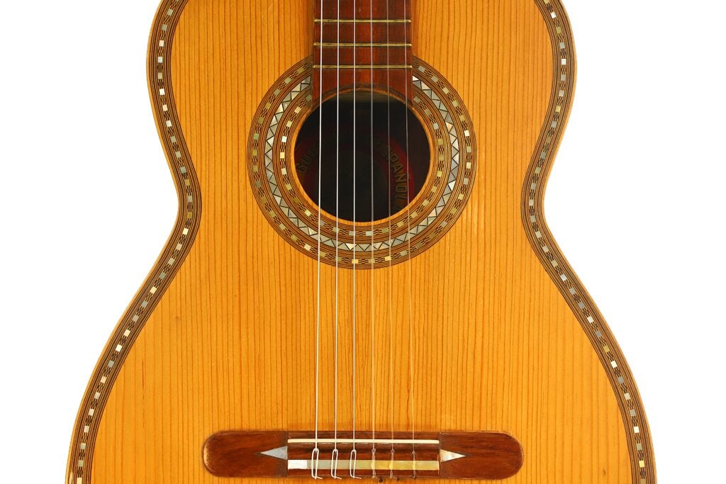 Nahaufnahme des Korpus einer „Klassische Gitarre“ von Salvador Ibanez aus dem Jahr 1920, die den edlen Holzfinish, die das Schallloch umgebende Zierrosette, den Steg und die sechs Saiten zeigt. Die Gitarre weist an den Rändern aufwendige Einlegearbeiten auf. Nahaufnahme des Korpus einer „Klassische Gitarre“ von Salvador Ibanez aus dem Jahr 1920, die den edlen Holzfinish, die das Schallloch umgebende Zierrosette, den Steg und die sechs Saiten zeigt. Die Gitarre weist an den Rändern aufwendige Einlegearbeiten auf.