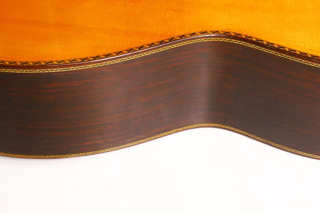 Nahaufnahme der gewölbten Seite einer klassischen Gitarre „Masaru Kohno Modell 50, 1979“, die die Holzmaserung und die Zierintarsien an den Kanten hervorhebt, vor weißem Hintergrund. Nahaufnahme der gewölbten Seite einer klassischen Gitarre „Masaru Kohno Modell 50, 1979“, die die Holzmaserung und die Zierintarsien an den Kanten hervorhebt, vor weißem Hintergrund.