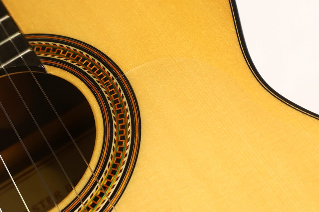 Nahaufnahme des Korpus einer Lester DeVoe 2020 Negra Flamenco-Gitarre, die einen Teil des Schalllochs, die Saiten und das dekorative Rosettenmuster auf hellem Holz zeigt. Nahaufnahme des Korpus einer Lester DeVoe 2020 Negra Flamenco-Gitarre, die einen Teil des Schalllochs, die Saiten und das dekorative Rosettenmuster auf hellem Holz zeigt.