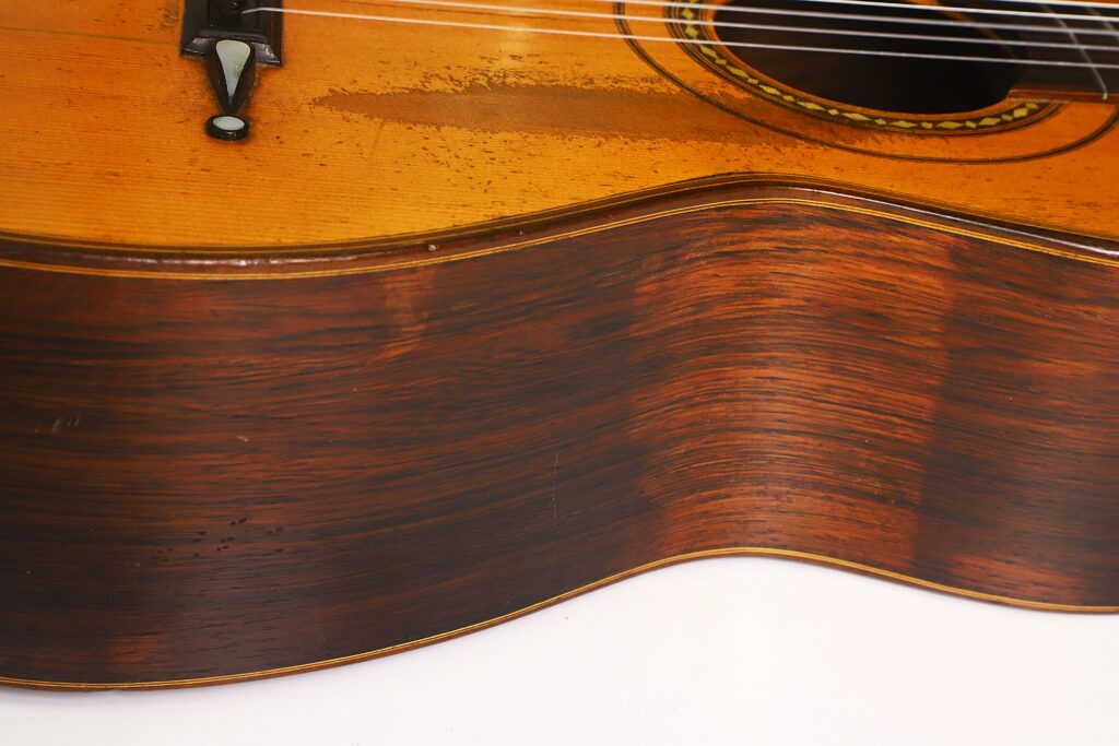 Nahaufnahme des Korpus einer „Romantikgitarre“ von Louis Panormo aus dem Jahr 1827, die die Holzmaserung, die Saiten, das Schallloch und einige Gebrauchsspuren auf der Oberfläche des Instruments zeigt. Nahaufnahme des Korpus einer „Romantikgitarre“ von Louis Panormo aus dem Jahr 1827, die die Holzmaserung, die Saiten, das Schallloch und einige Gebrauchsspuren auf der Oberfläche des Instruments zeigt.